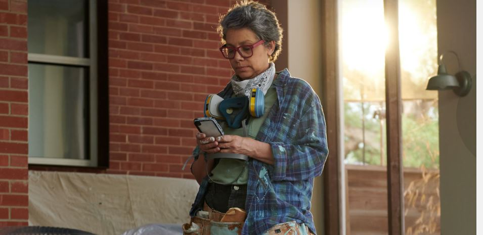 Painter looking at her phone. Wears red glasses, flannel shirt and tool belt, with respirator and bandana around neck. 