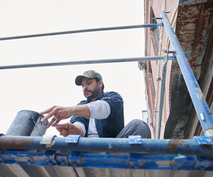 Person on a scaffold in front of a brick building dipping a brush into a can.