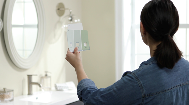 Person holding paint chips up to bathroom wall with sink, mirror and light fixture in background.